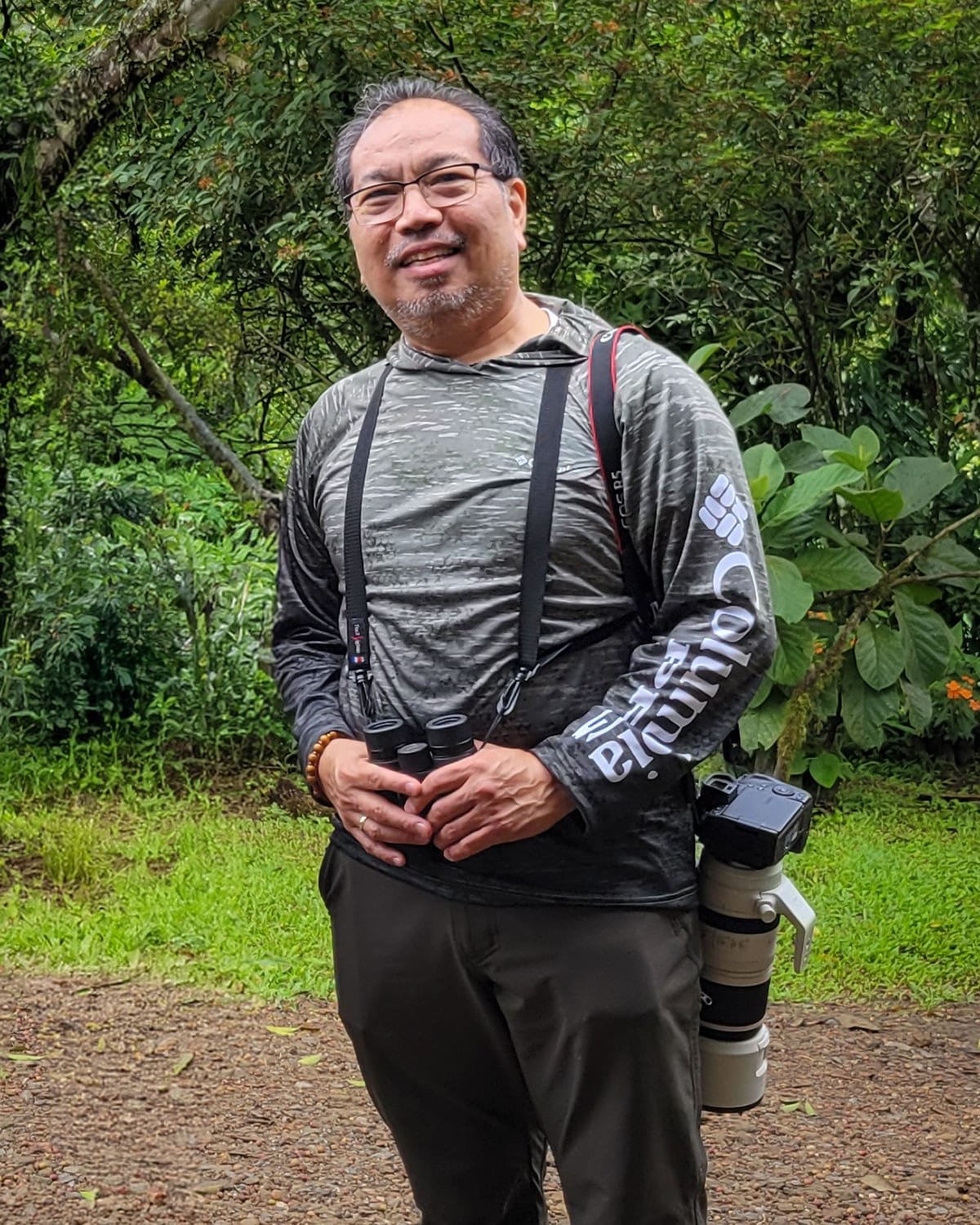 Eric Patdu standing on a forest trail in Costa Rica, binoculars on his chest and a telephoto lens at his side